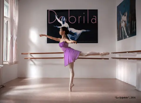 Balerina u ljubičastom kostimu u arabesci u sali škole, iza nje plakat sa imenom Dobrila